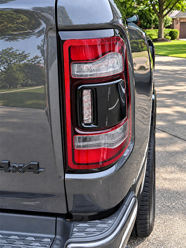 Close-up of the rear left side of a dark gray pickup truck, showing the taillight, rear quarter panel, tire, and a small portion of the 4x4 badge, with a driveway and trees in the background.