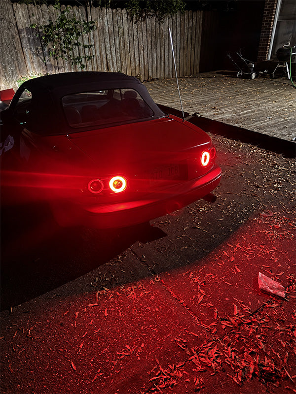 A red convertible car photographed at night in a backyard driveway, with its rear lights illuminated and casting a strong red glow onto the ground and deck.