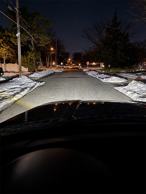 A view from inside a vehicle showing bright headlights illuminating a snowy residential street at night, with trees, utility poles, and a stop sign visible in the distance.