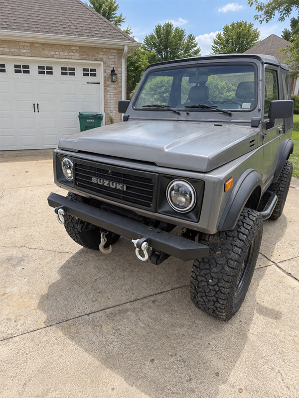 A silver-gray Suzuki Samurai off-road vehicle parked on a concrete driveway in front of a residential garage.