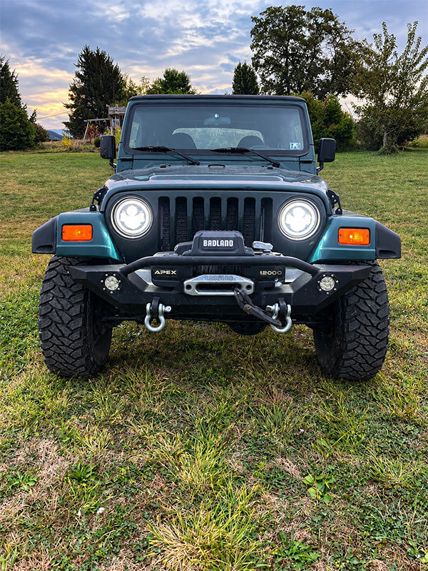 A front-facing view of a dark teal Jeep Wrangler parked on a grassy field, equipped with a heavy-duty black front bumper, winch, and off-road tires.