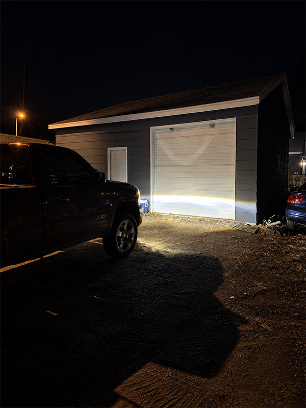 A black pickup truck parked in front of a gray garage door at night, with a bright beam of light shining onto the driveway. The scene is dark, emphasizing the light projection capability.