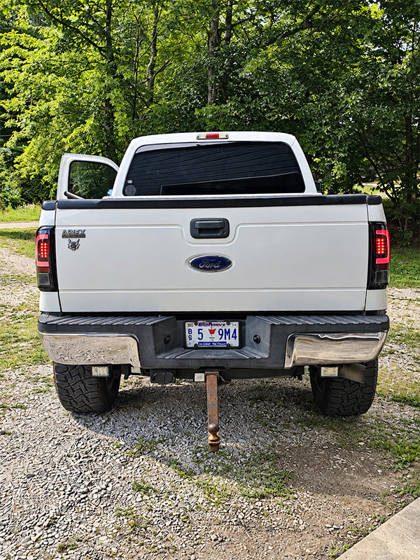 Rear view of a white Ford Ram pickup truck parked outdoors on a gravel surface with trees in the background. The driver-side door is open, and the tailgate, license plate, and hitch are visible.
