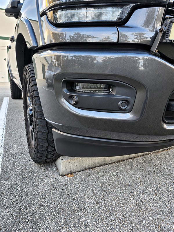Close-up of the front bumper and wheel area of a dark gray pickup truck parked on asphalt, showing the lower bumper, fog light housing, tire, and part of the headlight.