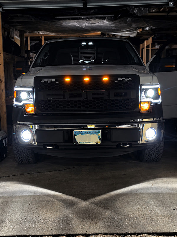 A front-facing view of a white Ford pickup truck parked inside a garage, with its headlights and fog lights turned on. The truck has a black grille and amber auxiliary lights mounted above the grille.