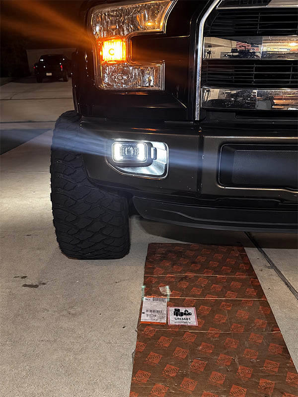 A close-up of a black truck's front bumper and headlight area at night, with a patterned brown and orange mat on the concrete ground in front of the bumper.