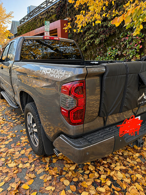 Rear three-quarter view of a dark gray Toyota Tundra 4x4 pickup truck parked outdoors on a road covered with fallen autumn leaves, with a black cover on the spare tire and a red paint splatter sticker on the rear bumper.