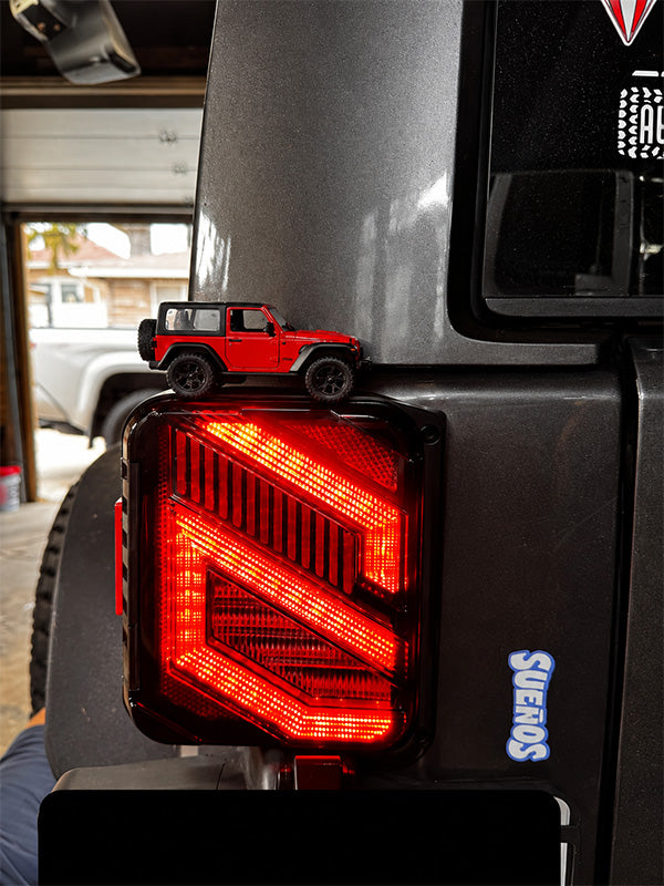 A red toy Jeep Wrangler sits on top of a black vehicle taillight with a geometric LED design. The photo is taken in a garage setting, with a gray vehicle and an open garage door visible in the background.