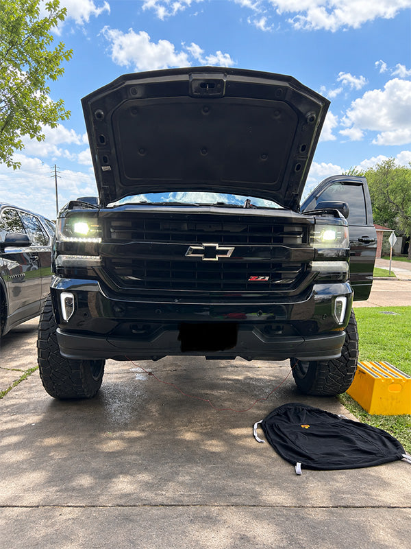 A black Chevrolet Silverado Z72 pickup truck with its hood open, parked on a concrete driveway outdoors under a blue sky with clouds. A black bag lies on the ground in front of the truck.