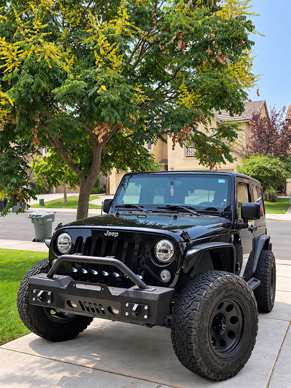 A black Jeep Wrangler parked on a concrete driveway in a suburban neighborhood, featuring a heavy-duty black front bumper and off-road tires.