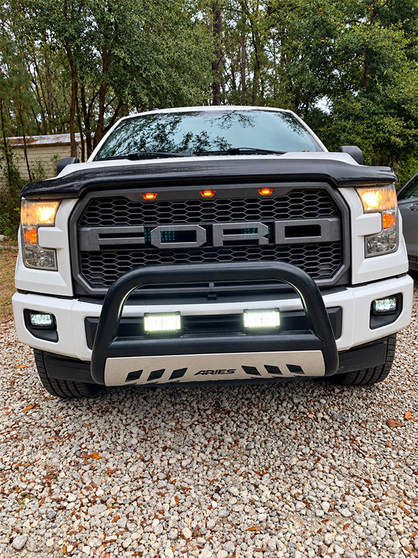 A white Ford pickup truck shown from the front, fitted with a black Aries bull bar and integrated LED light bars on the front bumper, parked on a gravel surface with trees in the background.