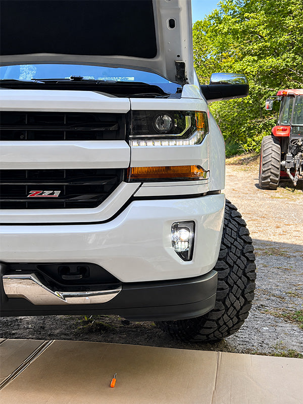 Close-up of a white pickup truck with the hood open, featuring an illuminated LED headlight and fog light, showcasing the lighting design and brightness in an outdoor garage setting.
