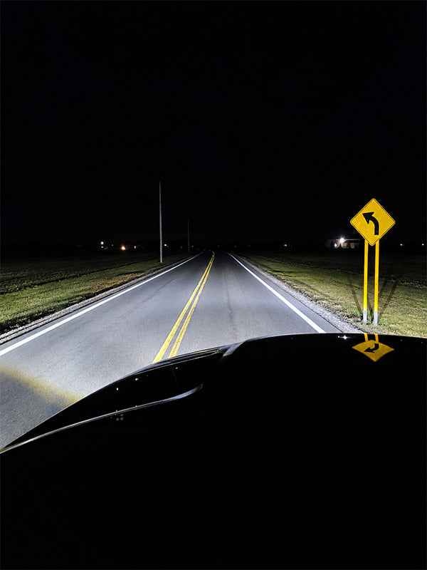 A nighttime front-facing view from inside a vehicle on a dark rural road, with the headlights illuminating the pavement and a yellow curved-road sign on the right side.