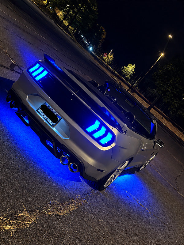 A gray sports car photographed at night on a street, with bright blue LED underglow and illuminated rear taillights casting blue light onto the pavement.