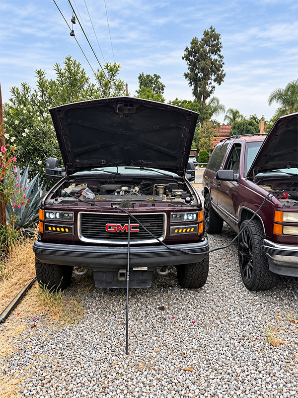 A dark maroon GMC SUV with its hood open, parked on a gravel driveway outdoors next to another vehicle under a blue sky with trees in the background.