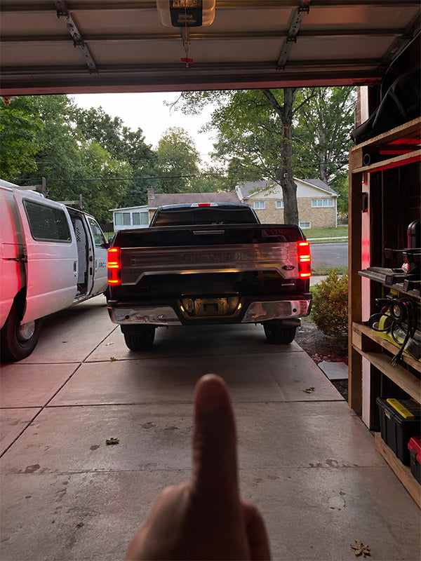 A black pickup truck is parked in a driveway inside a garage, viewed from behind as the garage door opens. A hand points toward the truck.