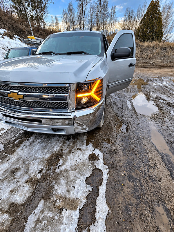 A silver Chevrolet pickup truck parked outdoors on a muddy, snow-dusted road with its front door open. The Chevrolet logo is visible on the grille.