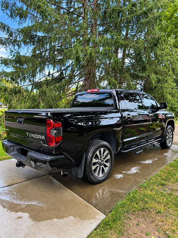 A black Toyota Tundra pickup truck parked on a wet concrete driveway with tall green trees and blue sky in the background.