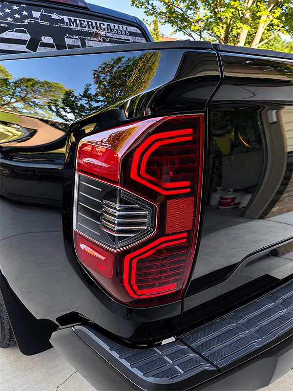 Close-up of the rear left taillight of a black pickup truck, showing the red LED light design and chrome reflector, with reflections of trees on the glossy black paint.