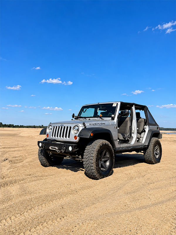 A silver Jeep Wrangler with its doors and windows removed, parked on a sandy beach under a bright blue sky.