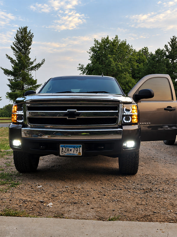 A front-facing view of a dark metallic Chevrolet pickup truck parked outdoors on a gravel surface, with the driver-side door open. Trees and a cloudy sky are visible in the background.