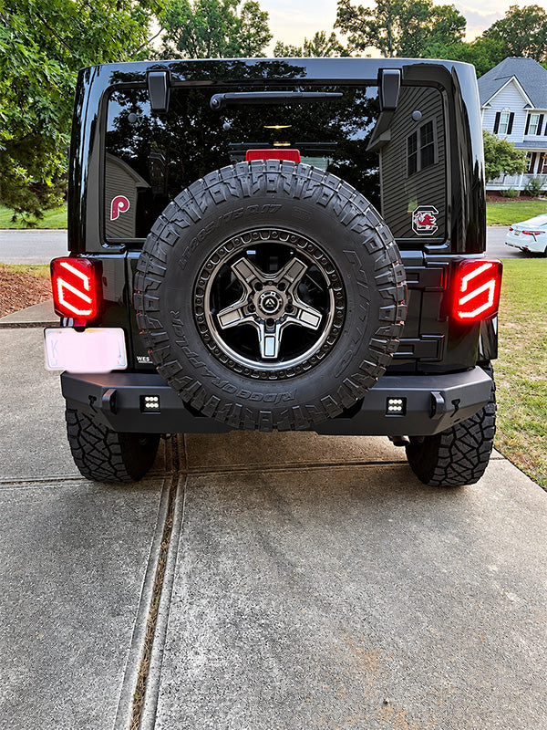 Rear view of a black off-road vehicle parked on a concrete driveway, featuring a spare tire mounted on the back, custom red LED tail lights, and a rear bumper with integrated lights.