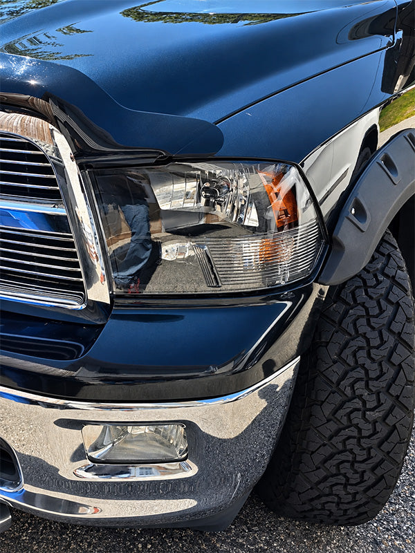 Close-up of the front left side of a dark blue Ram pickup truck, showing the headlight, chrome grille and bumper, and a black off-road tire.