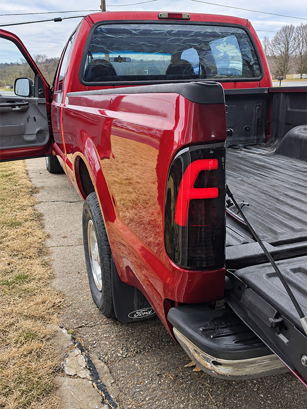 A red pickup truck parked outdoors on a paved surface beside grass, with the rear door open and the tailgate partially open. The product appears to be the truck itself rather than an auxiliary chart or diagram.