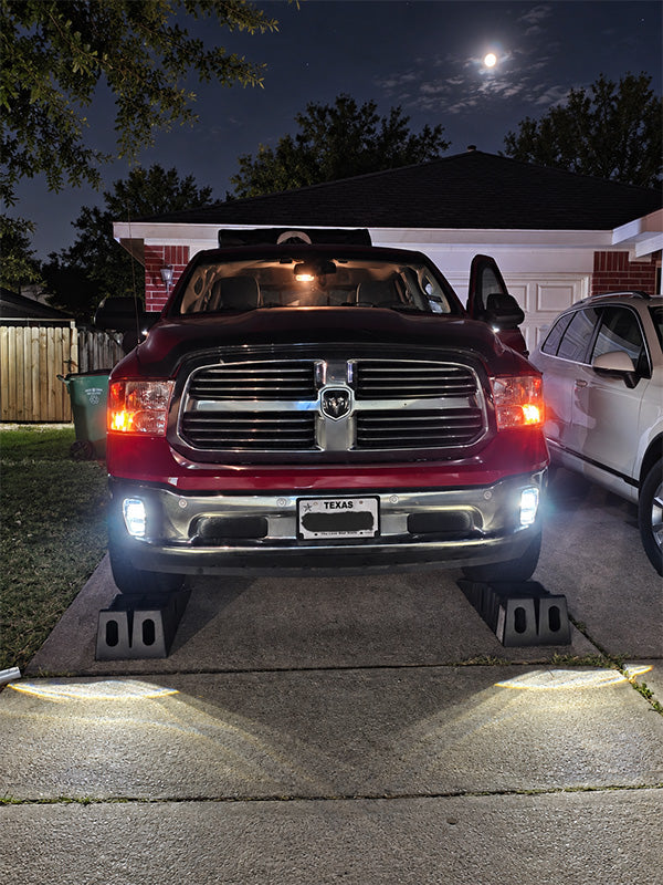 A red Ram pickup truck parked on a concrete driveway at night, with its headlights and fog lights turned on, illuminating the ground. The truck is positioned in front of a house with a visible moon in the sky.