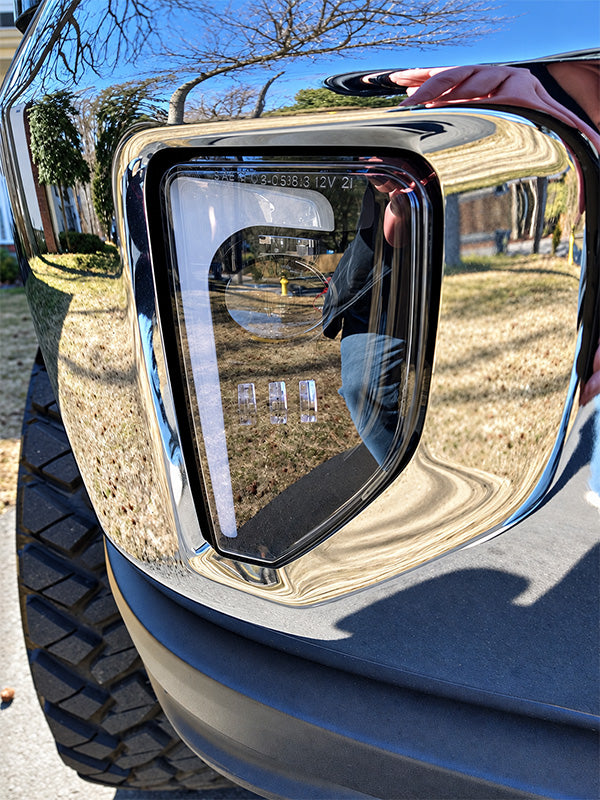 Close-up of a glossy black truck fender and wheel area with a highly reflective chrome bumper surround, showing outdoor reflections of trees, grass, and a person taking the photo.