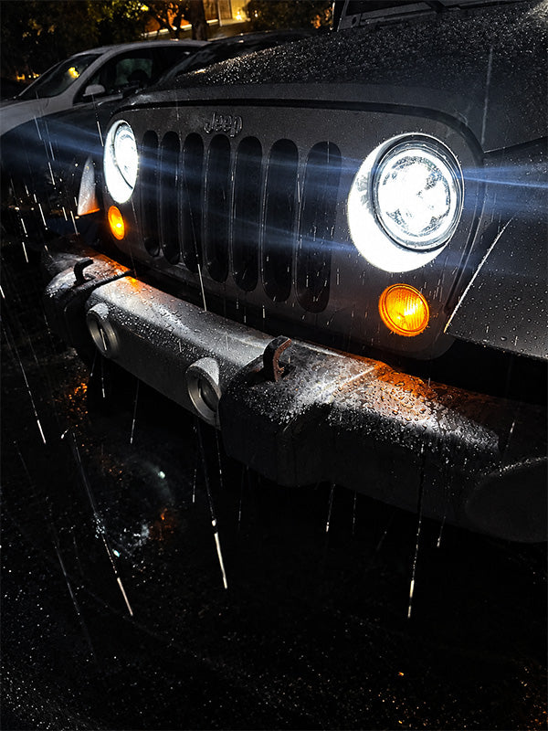 Close-up of the front of a black Jeep Wrangler parked in the rain at night, with headlights on, raindrops on the body, and orange turn signals glowing.