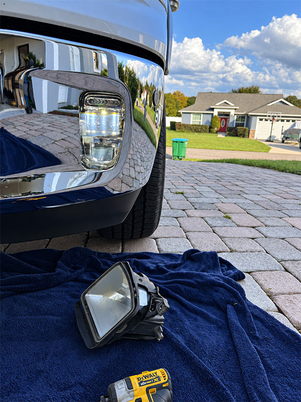 A chrome truck bumper headlight assembly is shown outdoors on a driveway with a blue towel and a yellow DeWalt cordless drill in the foreground, with a suburban house in the background.